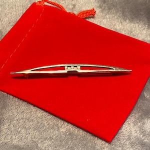 Puede incluir: Un broche plateado con un diseño estilizado, apoyado sobre una vibrante bolsa de terciopelo rojo. El broche tiene una forma delgada y curvada con extremos puntiagudos y un detalle central. El fondo es una tela gris suave.