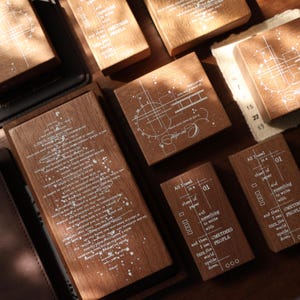 May include: A collection of rectangular wooden stamps featuring white printed text and geometric line designs. The stamps display various phrases including 'All I need is a sheet of paper and something to write with and then I can turn the world upside down' and 'One day it will all make sense'.