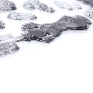 May include: Clear, transparent stamps of various shapes and sizes are scattered across a white surface. The stamps include a dress, a safety pin, and other decorative elements. The stamps are made of a clear material, allowing for easy alignment when stamping.