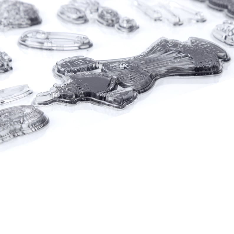 May include: Clear, transparent stamps of various shapes and sizes are scattered across a white surface. The stamps include a dress, a safety pin, and other decorative elements. The stamps are made of a clear material, allowing for easy alignment when stamping.