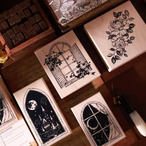 May include: A collection of rectangular wooden rubber stamps featuring black ink illustrations of a floral bouquet, an open window with vines, a castle under a moonlit sky, and a crescent moon seen through a window, alongside a box of letter stamps.