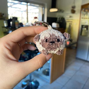 Peut inclure: Un petit porte-clés en peluche en forme d'animal rond, marron clair et blanc, avec de petites oreilles et un visage noir. Le porte-clés est attaché à un anneau en métal argenté.