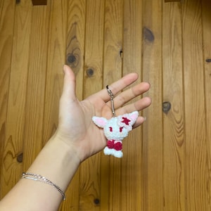 Peut inclure: Un porte-clés fait à la main en forme de lapin blanc au crochet, avec des détails roses sur les oreilles et un nœud papillon rouge. Le lapin a une étoile sur une oreille.