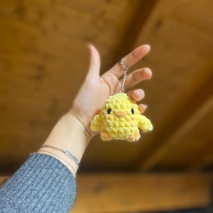 Peut inclure: Un porte-clés fait à la main en forme de poussin jaune au crochet, avec des yeux noirs et des détails orange. Le poussin est attaché à un porte-clés argenté. L'arrière-plan est un plafond en bois.