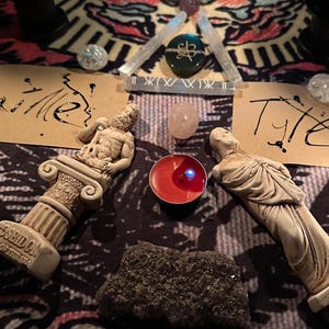 May include: A spiritual arrangement featuring two small statues, crystals, a lit red candle, and handwritten notes on a patterned cloth. The statues are beige, and the crystals include clear quartz and a pink stone. The candle emits a blue light.