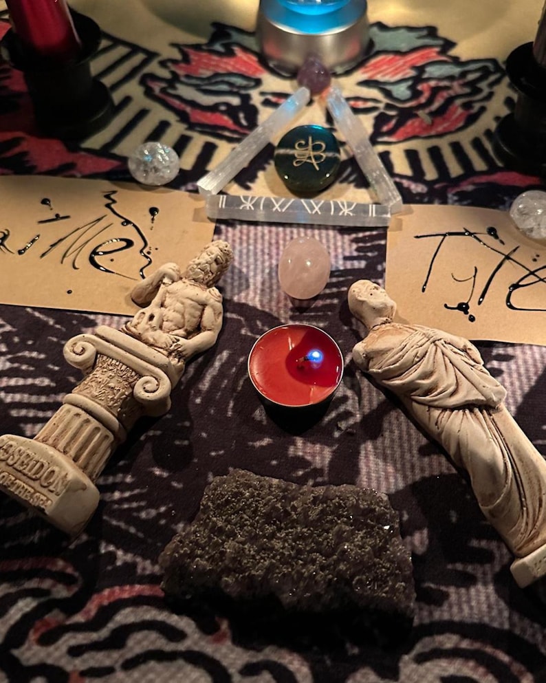 May include: A spiritual arrangement featuring two small statues, crystals, a lit red candle, and handwritten notes on a patterned cloth. The statues are beige, and the crystals include clear quartz and a pink stone. The candle emits a blue light.