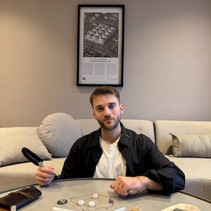 May include: A collection of spiritual items on a gray table, including a notebook, a feather pen, crystals, a necklace, and sage bundles. A framed black and white print hangs on the wall behind a person.
