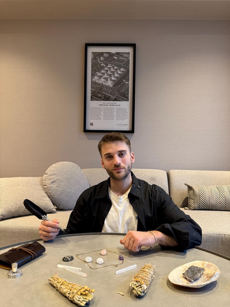 May include: A collection of spiritual items on a gray table, including a notebook, a feather pen, crystals, a necklace, and sage bundles. A framed black and white print hangs on the wall behind a person.