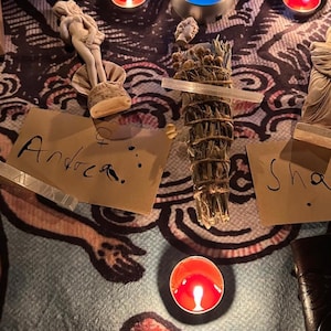 May include: A spiritual arrangement featuring a burning red candle, a bundle of sage, and small statues. The word "Andrea" is handwritten on a piece of paper. The scene is set on a patterned cloth, with additional candles and a blue light.