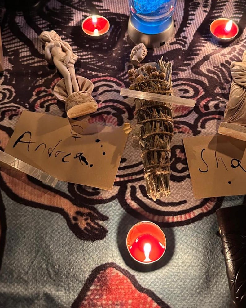 May include: A spiritual arrangement featuring a burning red candle, a bundle of sage, and small statues. The word "Andrea" is handwritten on a piece of paper. The scene is set on a patterned cloth, with additional candles and a blue light.