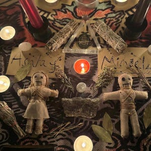May include: An overhead shot of a spiritual altar with candles, sage bundles, and voodoo dolls. The scene includes lit candles, dried herbs, and two dolls with pins. The names "Ashla" and "Michael" are written on paper.