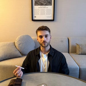 May include: A person seated at a round table with a leather-bound journal, a necklace with gemstones, and a white object. The person is wearing a black shirt over a white t-shirt. A framed poster is on the wall behind the person.
