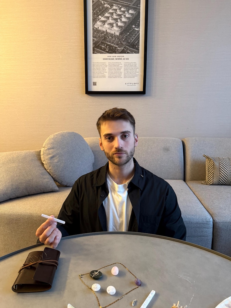 May include: A person seated at a round table with a leather-bound journal, a necklace with gemstones, and a white object. The person is wearing a black shirt over a white t-shirt. A framed poster is on the wall behind the person.