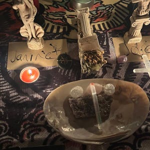 May include: A spiritual arrangement featuring a lit candle, a seashell bowl with crystals and a stone, and small statues. The word "Janire" is handwritten on a card. The background is a patterned cloth.