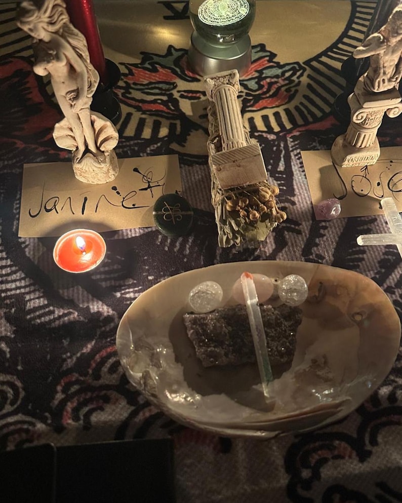 May include: A spiritual arrangement featuring a lit candle, a seashell bowl with crystals and a stone, and small statues. The word "Janire" is handwritten on a card. The background is a patterned cloth.