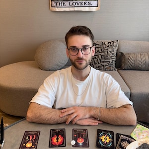 May include: A person wearing glasses and a white t-shirt sits at a table with tarot cards. The cards are black with colorful illustrations. A banner reading "THE LOVERS" hangs in the background. Sage bundles and crystals are also on the table.