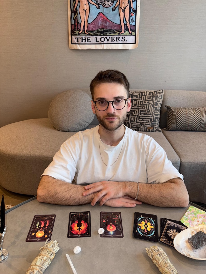 May include: A person wearing glasses and a white t-shirt sits at a table with tarot cards. The cards are black with colorful illustrations. A banner reading "THE LOVERS" hangs in the background. Sage bundles and crystals are also on the table.