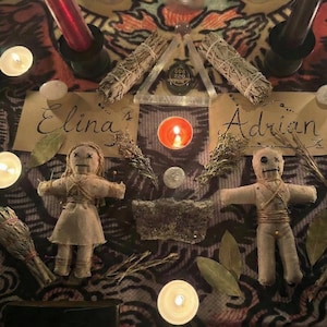 May include: An overhead shot of a spiritual altar with candles, sage bundles, and two voodoo dolls. The names "Elina" and "Adrian" are written on parchment. A triangular glass piece with text is centered above a lit candle.