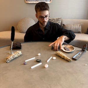 May include: A round, gray table displays various spiritual items. These include bundles of sage, crystals, a small dish, and an incense holder. A tarot card reading is in progress, with the card "The Lovers" visible in the background.