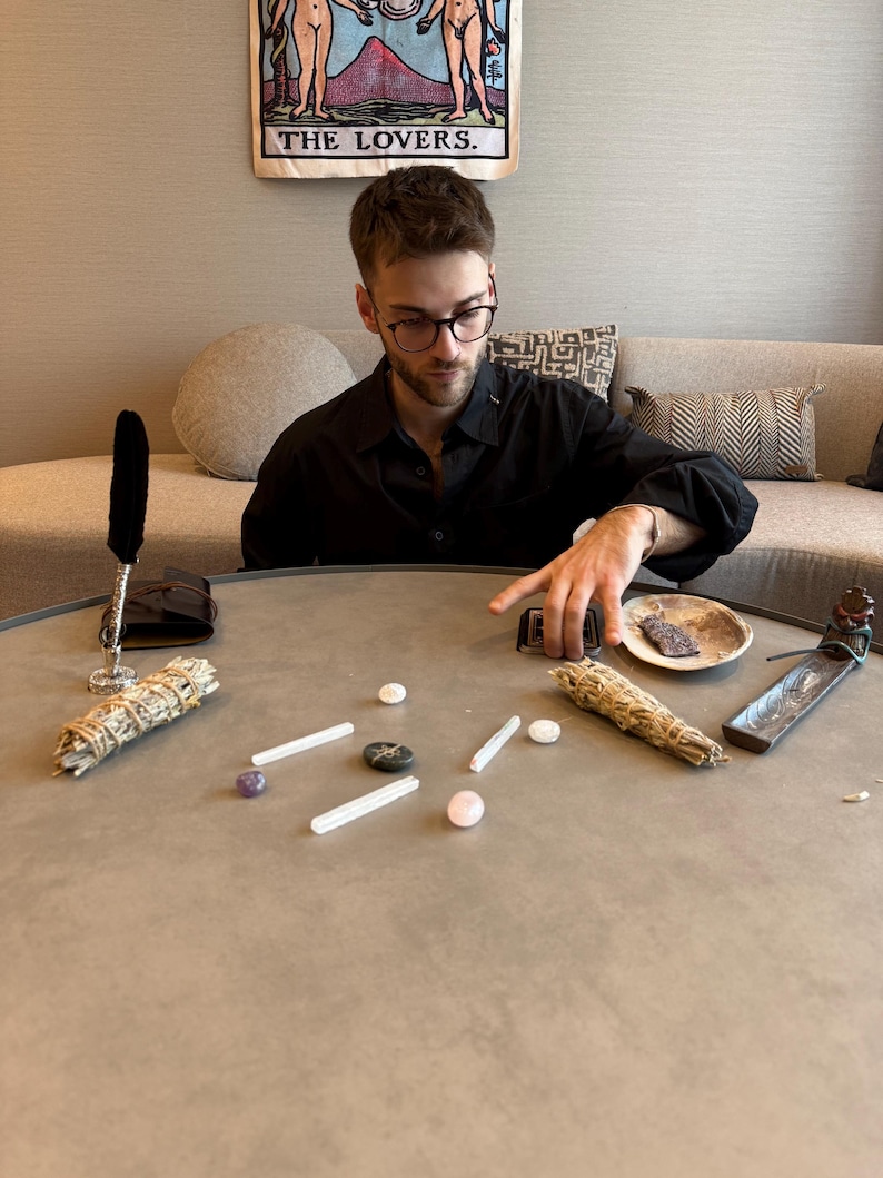 May include: A round, gray table displays various spiritual items. These include bundles of sage, crystals, a small dish, and an incense holder. A tarot card reading is in progress, with the card "The Lovers" visible in the background.