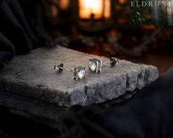 May include: A pair of square stud earrings with a clear, sparkling center stone. The earrings have a dark, decorative border and are set on a gray stone surface. The background features a blurred warm light and the word "ELDRUNE".