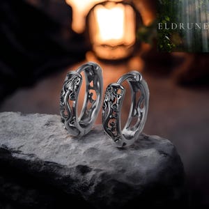 May include: A pair of silver-toned hoop earrings featuring an intricate, embossed dragon motif along the outer surface. The earrings have a hinged closure mechanism and a polished finish, showcasing detailed scales and serpentine body shapes.