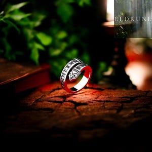 May include: A silver-colored ring with a red interior. The ring features engraved symbols and text around the band. The ring is displayed on a textured, brown surface with a blurred green background. The word "ELDRUNE" is visible in the upper right corner.