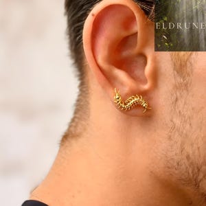 Può includere: Un orecchino a forma di millepiedi color oro è indossato sull'orecchio. L'orecchino è un design dettagliato e tridimensionale. L'orecchino è attaccato all'orecchio. Lo sfondo è sfocato, con la parola ELDRUNE visibile in alto a destra.