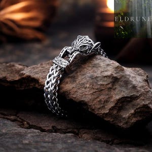 May include: A silver-toned metal bracelet featuring a detailed wolf head terminal with intricate knotwork patterns. The chain consists of a woven, interlocking link design. The piece includes the text ELDRUNE in the upper right corner.