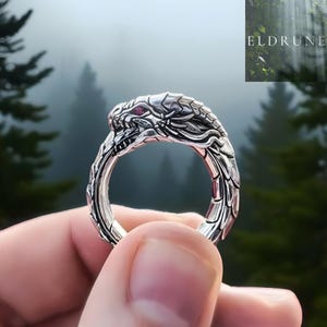 Op de afbeelding: Een zilveren ring in de vorm van een opgerolde draak met gedetailleerde schubben en een kleine rode edelsteen als oog. De ring heeft overal ingewikkelde metaalstructuren. De tekst ELDRUNE is zichtbaar in de rechterbovenhoek.