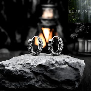 Puede incluir: Dos pendientes de aro plateados con un diseño ornamental simétrico. Los pendientes se exhiben sobre una roca gris texturizada. El fondo es oscuro, con una fuente de luz borrosa y la palabra "ELDRUNE" visible.