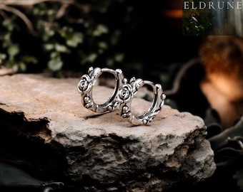 May include: Silver-coloured hoop earrings with a floral design. Each earring has rose-shaped details. The earrings are displayed on a textured, grey stone surface. The word "ELDRUNE" is visible in the background.