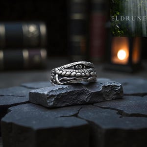 Può includere: Un anello d'argento a forma di testa di serpente, con squame dettagliate e un occhio. L'anello è appoggiato su una superficie scura e strutturata, con libri sfocati sullo sfondo. Il design dell'anello è intricato e accattivante.