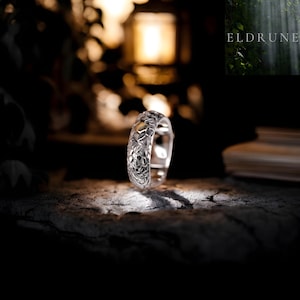 Peut inclure: Une bague en argent avec un motif texturé complexe. La bague présente un motif répétitif sur le bracelet et une petite pierre de couleur claire. Le mot "ELDRUNE" est visible en arrière-plan.