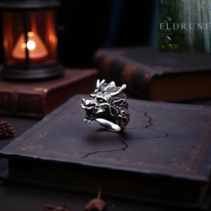 Könnte beinhalten: Ein silberner Drachenring mit detailliertem Drachenkopf-Design. Die filigranen Merkmale des Rings werden vor einem dunklen Hintergrund hervorgehoben. Der Ring ist ein Statement-Schmuckstück.