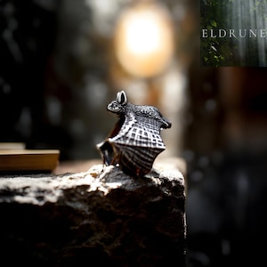 May include: A detailed, silver-toned bat figurine with textured wings and body, perched on a rough stone surface. The bat's wings are spread, and the design includes intricate patterns. The background is blurred, with a soft light source and the text "ELDRUNE".