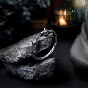 May include: A silver-toned bracelet with wolf head details. The bracelet features a twisted cable design and is displayed on a gray rock. The word "ELDRUNE" is visible in the background.