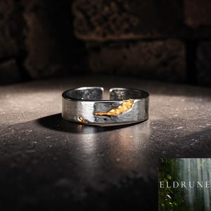 May include: A silver-toned open band ring featuring a textured, brushed metal finish and irregular, recessed gold-colored accents. The ring has a visible gap at the back, allowing for size adjustment. The word ELDRUNE is printed in the bottom corner.