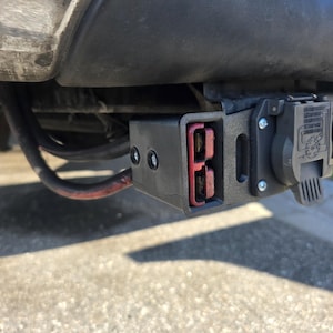 May include: A black trailer hitch electrical connector assembly mounted under a vehicle bumper. The unit features a rectangular housing with a dual-port red power connector and a circular seven-way trailer plug with a hinged cover and wiring diagram.