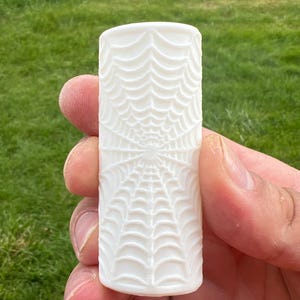 May include: A cylindrical white object featuring a raised, embossed spider web pattern that covers the entire surface. The design consists of radiating lines connected by concentric curved segments, creating a detailed, textured web motif.