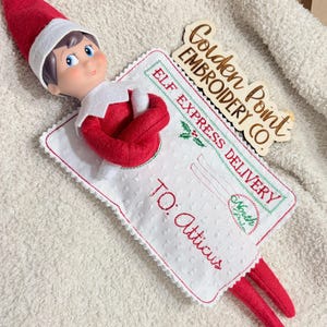 May include: A plush elf doll with a red hat and suit attached to a white rectangular fabric envelope. The envelope features red embroidered text reading ELF EXPRESS DELIVERY and TO: Atticus, with a North Pole stamp and a wooden sign nearby.