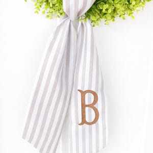 May include: A decorative door hanger featuring a green wreath with a striped ribbon. The ribbon is white and tan with a large embroidered brown letter "B". The wreath is made of small green leaves.