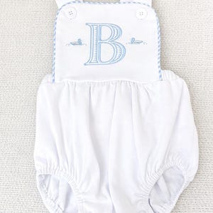 May include: White baby romper with blue embroidered details. The romper features a large blue letter "B" and two small duck illustrations. The straps and neckline are trimmed with blue and white striped fabric. The romper has snap closures.