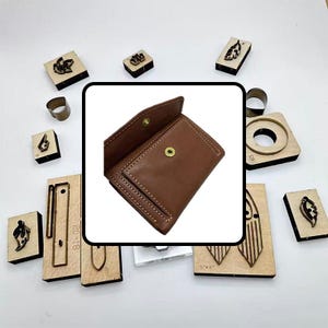 Puede incluir: Una cartera de cuero marrón con una solapa abierta, rodeada de varias herramientas y sellos de madera. Las herramientas incluyen formas de hojas y geométricas, y un anillo de metal. La cartera tiene un cierre de botón a presión.