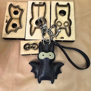 May include: A black leather bat-shaped keychain with a silver metal lobster clasp and a wrist strap, positioned in front of four wooden cutting dies with metal blades used for crafting the bat shape and its individual components.