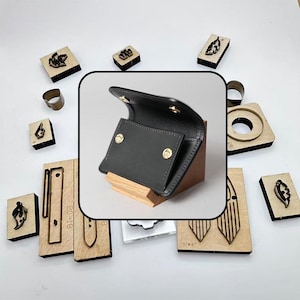 May include: A black leather wallet with gold-tone snap buttons, displayed on a wooden stand, surrounded by various metal leather-cutting dies of different shapes and sizes arranged on a plain white surface.
