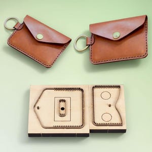 Może przedstawiać: Dwa brązowe skórzane portmonetki z kółkami na klucze i mosiężnymi zatrzaskami. Poniżej pokazano drewnianą formę do wykrawania o skomplikowanych kształtach do tworzenia wyrobów skórzanych.