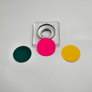 Può includere: Vista dall'alto di un quadrato in acrilico trasparente con un taglierino circolare in metallo. Tre cerchi colorati lo circondano: verde scuro, rosa acceso e giallo. Gli oggetti sono su una superficie bianca.