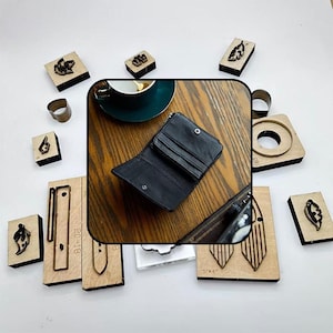 May include: An open black leather wallet with multiple compartments sits on a wooden surface, surrounded by various wooden craft tools and stamps. The tools include leaf and geometric shapes, and a metal ring.