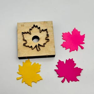Könnte beinhalten: Ein hölzernes Stanzwerkzeug mit Ahornblattform und der Nummer 55. Drei farbige Ahornblatt-Ausschnitte in Gelb, Pink und Magenta befinden sich neben dem Stanzwerkzeug. Das Stanzwerkzeug hat ein kreisförmiges Loch in der Mitte.